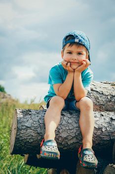 A boy in a blue baseball cap and denim shorts sits on a log and looks thoughtfully