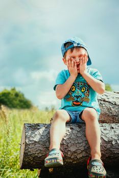 A boy in a blue baseball cap and short shorts sits on a log playing and twisting his face