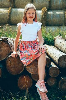 A young girl with long blonde hair sits smiling on a log in a pink dress