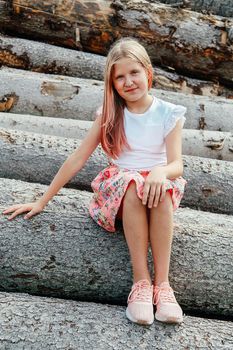 Blonde girl with long flowing hair sits smiling on a log in a pink dress
