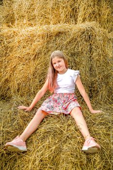 A schoolgirl with long blonde hair sits like a doll in a pink dress on a bale of straw on a hot summer day
