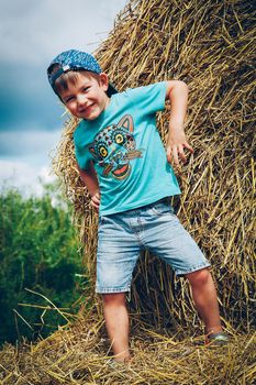 A boy in a blue baseball cap and shorts having fun and playing on round bales of straw on a hot summer day