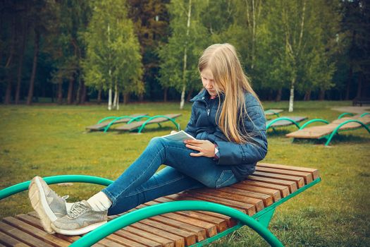 Girl with blond hair writing a letter on a wooden chaise longue in the spring