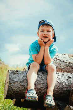 A little boy in a blue baseball cap and shorts sits on a log and peers into the distance