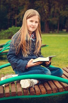 Blonde school-age girl sitting on a bench in the Park and reading a book