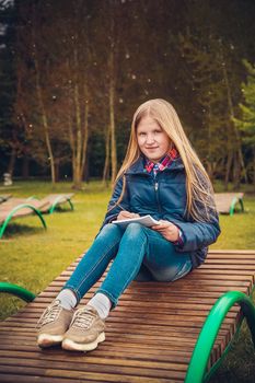 Blonde girl writes a letter in the spring sitting on a wooden chaise longue in the Park