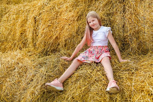A blonde girl with long hair in a pink dress sits on a bale of straw on a hot summer day like a doll