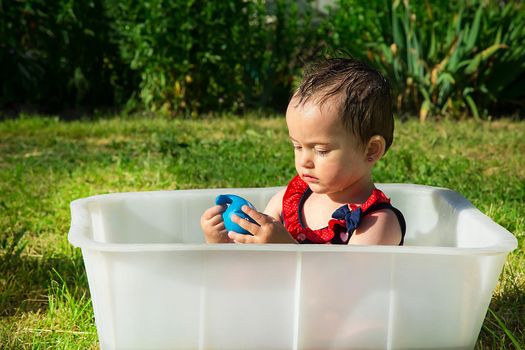 On a hot summer day, a very serious year-and-a-half girl plays with a blue rubber toy in a white bath