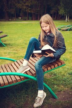 Girl with blond school-age hair sitting on a wooden chaise longue in the Park and reading a book