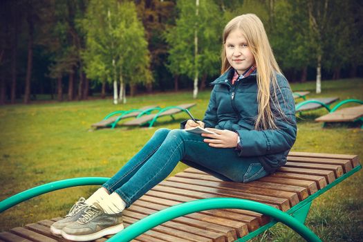 Blonde girl writes notes sitting on a wooden chaise longue in the Park in nature