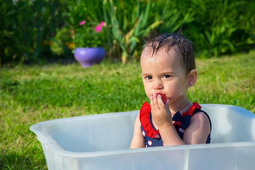 A little girl in a swimsuit sitting in a white bath and eating strawberries