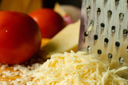 Stock photo of pile of shredded cheese and grater with couple of red tomatoes in the blurred background in close-up.