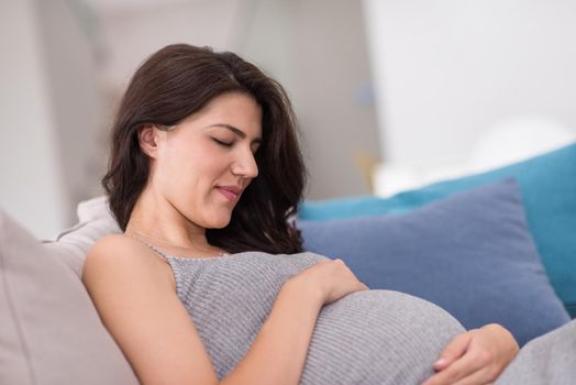 young happy pregnant woman keeping hand on belly while sitting on sofa at home