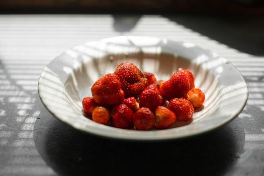 picture of juicy fresh ripe red strawberries in a white ceramic plate on the table under bright sunlight in a village