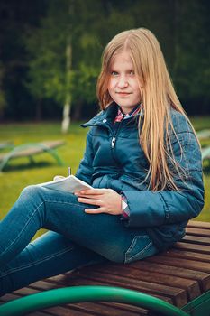 Blonde girl sitting on a wooden chaise longue and writing a letter