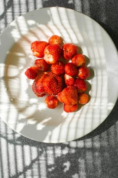 picture of juicy fresh ripe red strawberries in a white ceramic plate on the table under bright sunlight in a village