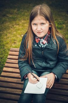 A school-age girl in the spring sitting on a wooden chaise longue in a forested area writes a letter