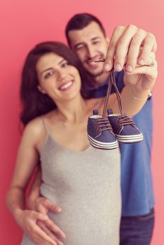 young  pregnant couple holding newborn baby shoes isolated on red background,family and parenthood concept