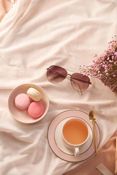 Cup of tea, macarons cake, glasses and flowers on fabric background