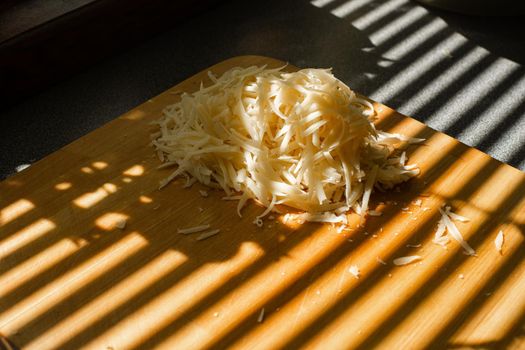 Stock photo of pile of fresh yellow grated cheese on wooden cutting board on kitchen table covered with blinds shade. Stripes from blinds on the window casting over board with shredded cheese.