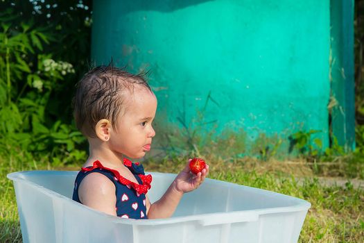 A little girl in a swimsuit sits in a bath and eats ripe red strawberries