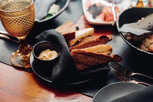 Stock photo of slices of white and brown bread on black napkin with a sauce in black saucer on black plate on served table in restaurant.