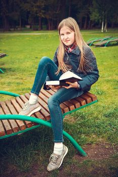 School-age girl sitting on a wooden chaise longue in the Park and reading a book