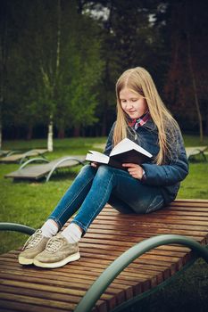 Schoolgirl with blond hair sitting on a wooden chaise longue in the Park and reading a book