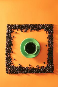 Green cup of coffee in the middle of quare shape coffee beans on orange background
