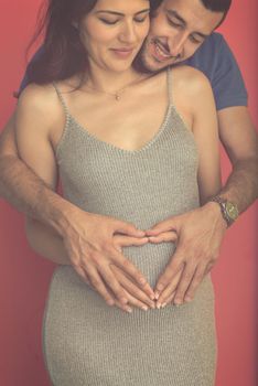 Happy young couple showing heart with their hands on belly of pregnant wife isolated on red background