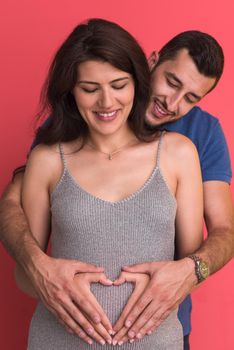 Happy young couple showing heart with their hands on belly of pregnant wife isolated on red background