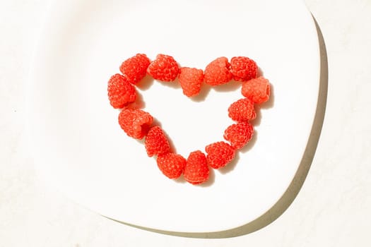 Top view of fresh and succulent raspberries in shape of heart on white ceramic dish. Concept of love. Juicy raspberries dessert.