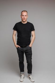 picture of attractive man wearing a black t-shirt and jeans stands with hands in pockets isolated on grey background