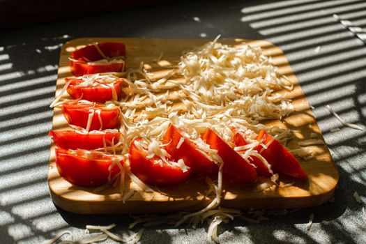 Stock photo in close up of sliced tomatoes with shredded cheese on wooden board in sunlight. Blinds shadow over the table. Cooking process.