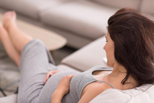 young happy pregnant woman keeping hand on belly while sitting on sofa at home