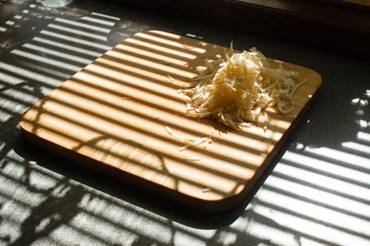 Stock photo of pile of fresh yellow grated cheese on wooden cutting board on kitchen table covered with blinds shade. Stripes from blinds on the window casting over board with shredded cheese.