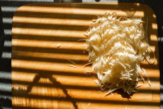 Stock photo of pile of fresh yellow grated cheese on wooden cutting board on kitchen table covered with blinds shade. Stripes from blinds on the window casting over board with shredded cheese.