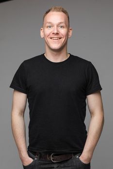 Studio portrait of cheerful blond Caucasian man in black t-shirt and jeans holding hands in pockets and smiling at camera on grey background.