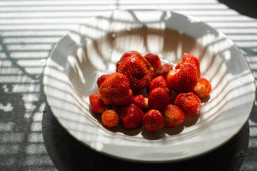 picture of juicy fresh ripe red strawberries in a white ceramic plate on the table under bright sunlight in a village