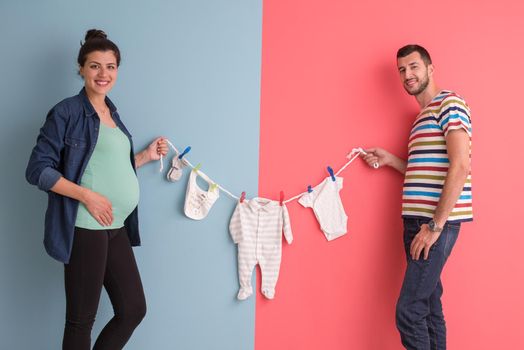 Beautiful pregnant woman and her husband expecting baby holding baby bodysuits and smiling over colorful background