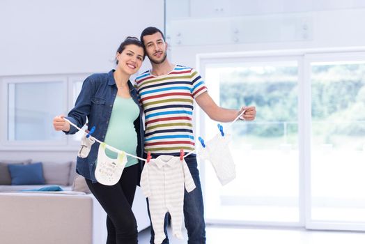 Beautiful pregnant woman and her husband expecting baby holding baby bodysuits and smiling at home