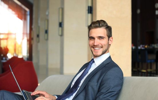 Young businessman working on laptop, sitting in hotel lobby waiting for someone