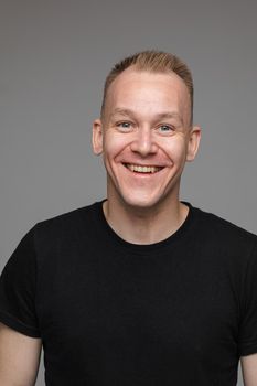 Studio portrait of adult man with short blond hair wearing casual black t-shirt smiling cheerfully at camera with friendly look. Cutout on grey background.