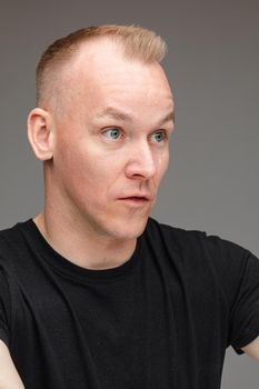 Portrait of astonished blond Caucasian man in black talking to someone showing irritation and bewilderment with cropped arms up on grey background.