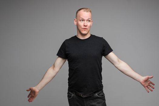 Studio portrait of handsome adult Caucasian man with short blond hair in black t-shirt and dark jeans looking away with puzzled face and outstretched arms.