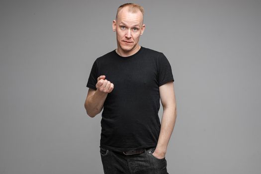 Studio portrait of a strict man with threatening face in black t-shirt and jeans showing fist at camera with other hand in pocket.