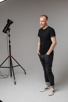 Full length portrait of stylish Caucasian man with short blond hair wearing casual black t-shirt, dark jeans and white sneakers standing with hands in pockets. Studio light in background.