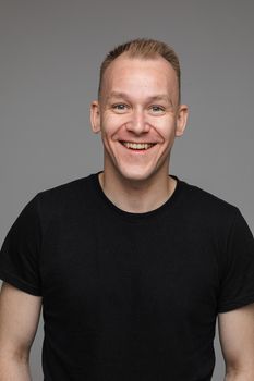 Studio portrait of adult man with short blond hair wearing casual black t-shirt smiling cheerfully at camera with friendly look. Cutout on grey background.