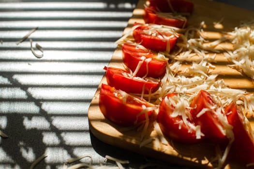 Stock photo in close up of sliced tomatoes with shredded cheese on wooden board in sunlight. Blinds shadow over the table. Cooking process.