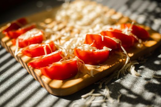 Stock photo in close up of sliced tomatoes with shredded cheese on wooden board in sunlight. Blinds shadow over the table. Cooking process.